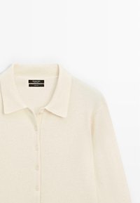 Cardigan en laine crème avec un col à revers, une fermeture à boutons et une texture douce. Étiquette visible à l'intérieur du col indiquant la marque et le matériau.