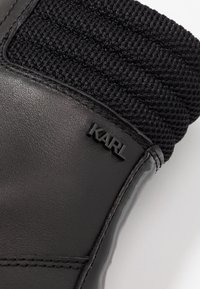 Bota de cuero negra con un puño de tela negra texturizada y elástica. Presenta un detalle del logo "KARL" elevado en el lateral. Acabado suave.