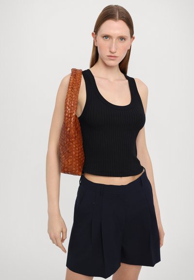 Schwarzes geripptes Tanktop, kurzes Design, kombiniert mit navyblauen, plissierten Shorts. Das Model hält eine gewebte, braune Ledertasche. Neutraler Hintergrund.