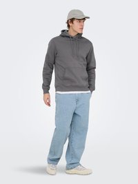 Jonge man met een lichtgrijze pet, donkergrijze hoodie, lichtblauwe spijkerbroek en beige sneakers, staand en naar rechts kijkend tegen een witte achtergrond.