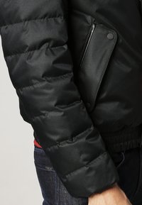 Chaqueta acolchada negra con textura lisa y brillante. Cuenta con un bolsillo con cremallera y puños acanalados, diseñada para ofrecer calidez y comodidad.