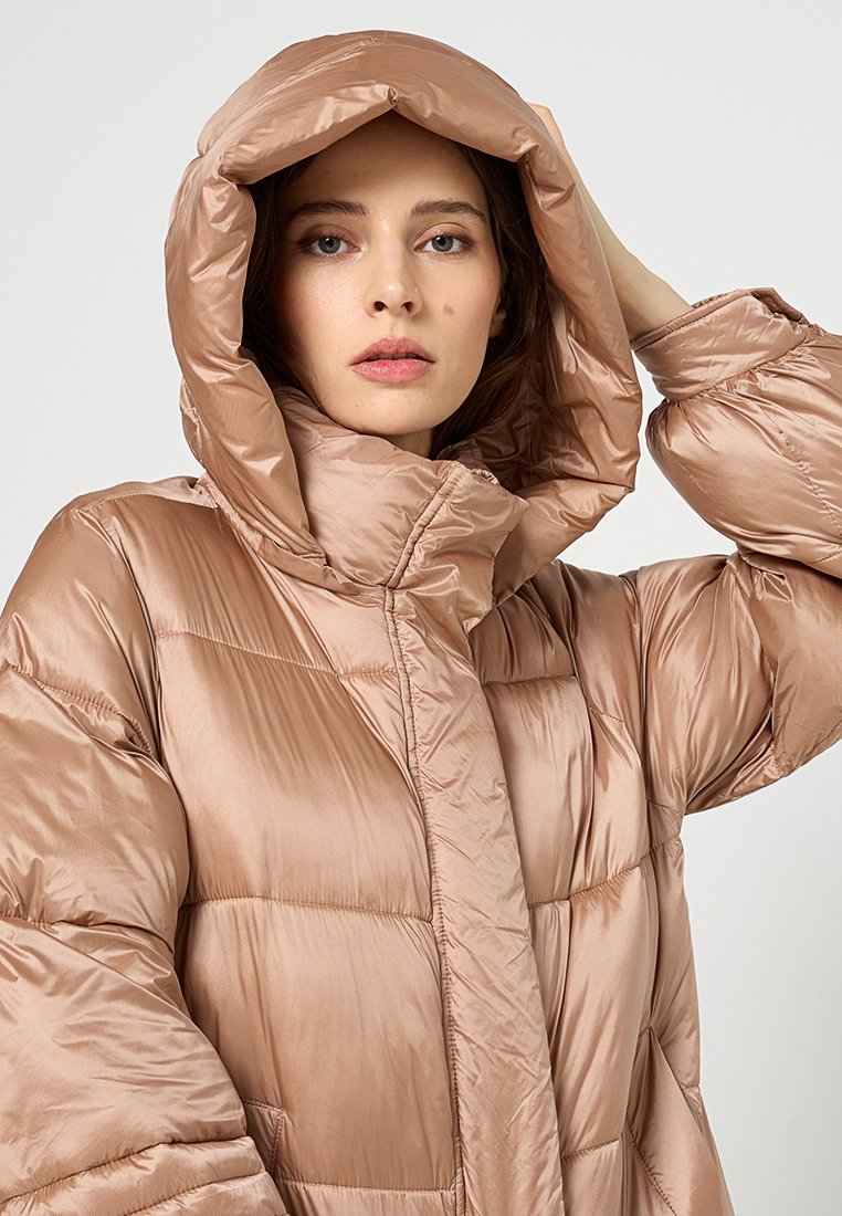 Kurtka puchowa w miękkim, błyszczącym beżowym materiale z dużym kapturem, dużym pikowaniem w romby oraz z zamkiem błyskawicznym z przodu.