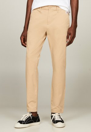 Chinos de algodón beige con corte slim, que cuentan con un cierre de botón, bolsillos laterales y un pequeño parche de logo en la cinturilla. Combinados con zapatillas negras.