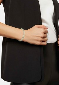 Pulsera de cuentas multicolores que presenta cuentas de color verde claro y transparentes, llevada en una mano sobre un fondo de chaleco oscuro y blusa blanca.