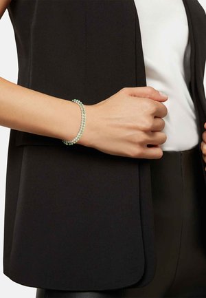 Bardzo kolorowa bransoletka z koralików, składająca się z jasnych zielonych i przezroczystych koralików, noszona na ręce na tle ciemnej kamizelki i białej bluzki.