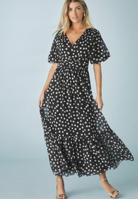 Rochie maxi cu buline negre, cu mâneci fluier și talie strânsă, cu un fustă în straturi. Material transparent cu o textură moale și o siluetă fluidă.