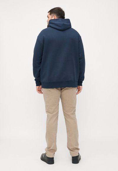 Hanorac cu glugă de culoare bleumarin, cu o croială lejeră, ce prezintă mâneci lungi, tivuri înfășurate și un buzunar tip cangur. Asortat cu pantaloni kaki și încălțăminte neagră.