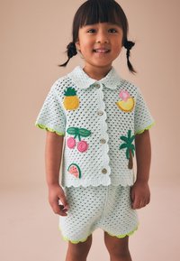 Niño pequeño sonriendo, vistiendo una camisa y pantalones cortos de crochet azul claro decorados con parches coloridos de frutas y palmeras.