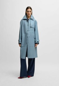 Light blue trench coat with a high collar, front buttons, side pockets, and oversized sleeves, paired with navy trousers and red shoes.