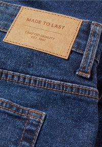 Denim fabric with a brown leather patch labeled "MADE TO LAST," featuring embossed text and stitched edges. Dark blue color.