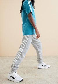 Turkusowa koszulka z krótkim rękawem, szare spodnie dresowe z białymi pasami i białe buty sportowe z czarnymi akcentami. Neutralne tło.