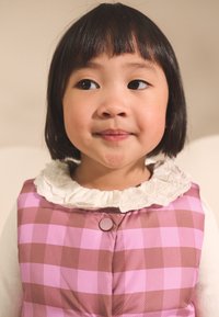 Pink og brun ternet dynejakkevest med en hvid flæsekkrave og knaplukning, båret af et barn med kort sort hår.