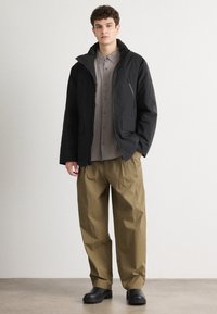 Giacca nera leggera con cappuccio, tasche con zip, abbinata a una camicia grigia con bottoni e pantaloni khaki larghi. Calzature nere completano il look.