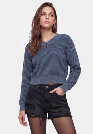 Ung kvinde iført en blå tekstureret langærmet sweater, sorte revnede denimshorts og sorte strømpebukser, står med den ene hånd i lommen.