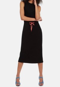 Schwarzes ärmelloses Midikleid mit einem taillierten Oberteil und einem lockeren Rock, ausgestattet mit einem roten Kordelzug in der Taille. Getragen mit blauen Sandaletten mit Riemchen.