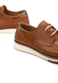 Scarpe in pelle marrone con una finitura liscia, dotate di una suola beige chiaro, design con lacci e cuciture decorative lungo i bordi.