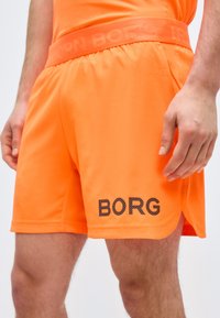 Κοντινό πλάνο ενός ατόμου που φοράει φωτεινά πορτοκαλί σορτς με την επιγραφή "BORG" σε μαύρο στο αριστερό πόδι και αντιστοιχία πορτοκαλί ζώνη μέσης.