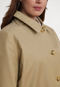 Μπεζ καμπαρντίνα με μυτερό γιακά, που διαθέτει μεγάλα διαφανή κουμπιά, λεία υφή και άνετη γραμμή. Κοντινή λήψη.