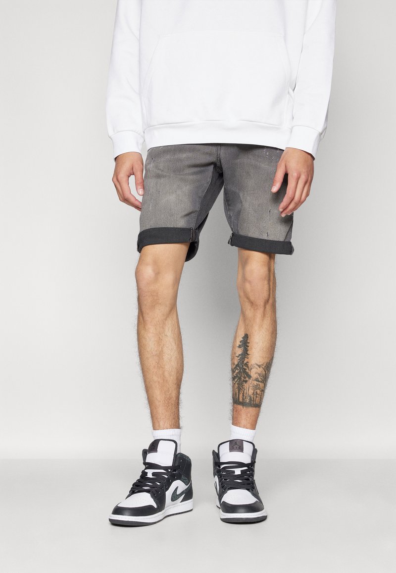 Shorts in denim grigio con orli arrotolati, abbinati a sneakers alte in bianco e nero. Presenta un tatuaggio visibile sulla parte bassa della gamba.
