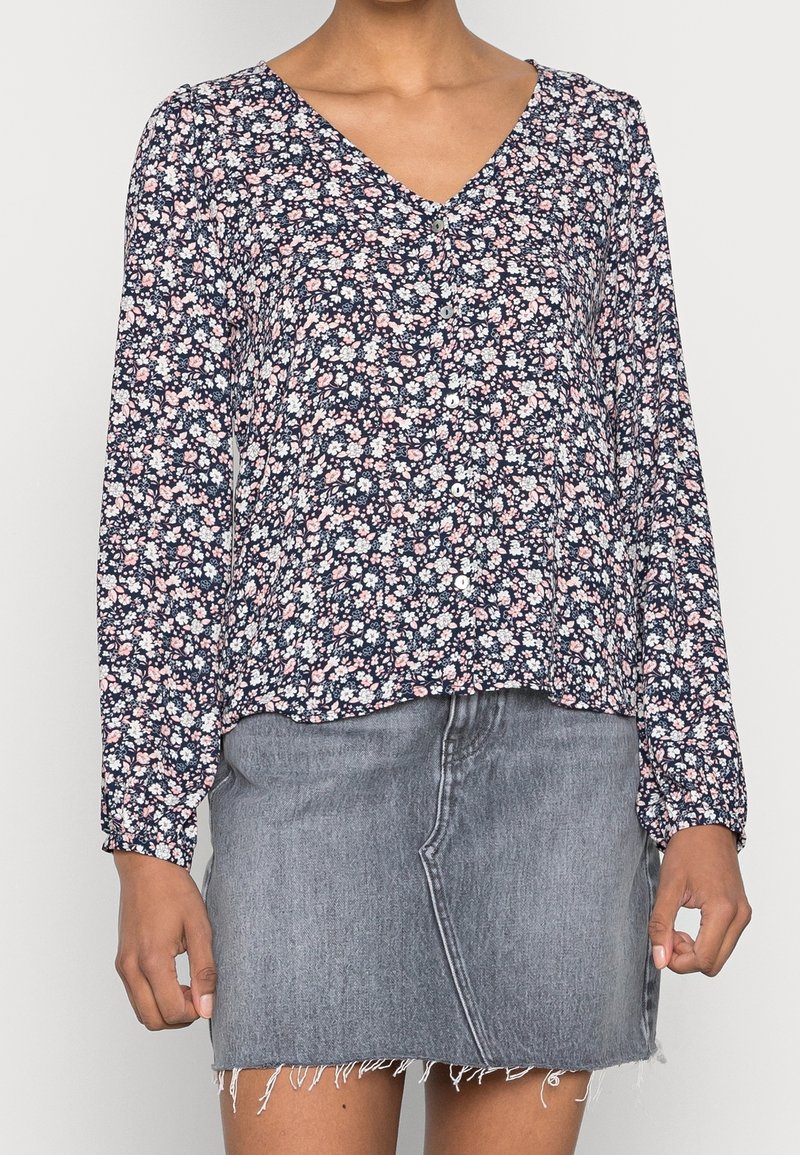 Blouse longue à manches longues à motifs floraux avec un fond bleu marine, des fleurs blanches et roses, un col en V et des détails de boutons. Associée à une jupe en jean grise.