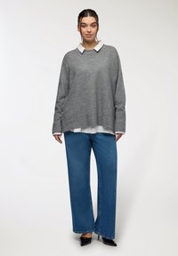 Maglione grigio a maglia con scollo rotondo, indossato sopra una camicia bianca con colletto. Pantaloni larghi in denim blu e tacchi neri.