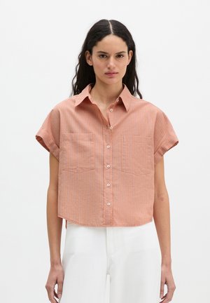 Kobieta ubrana w pomarańczową, krótką, zapinaną na guziki, krótką koszulę w pionowe paski oraz białe spodnie z wysokim stanem na jednolitym tle.