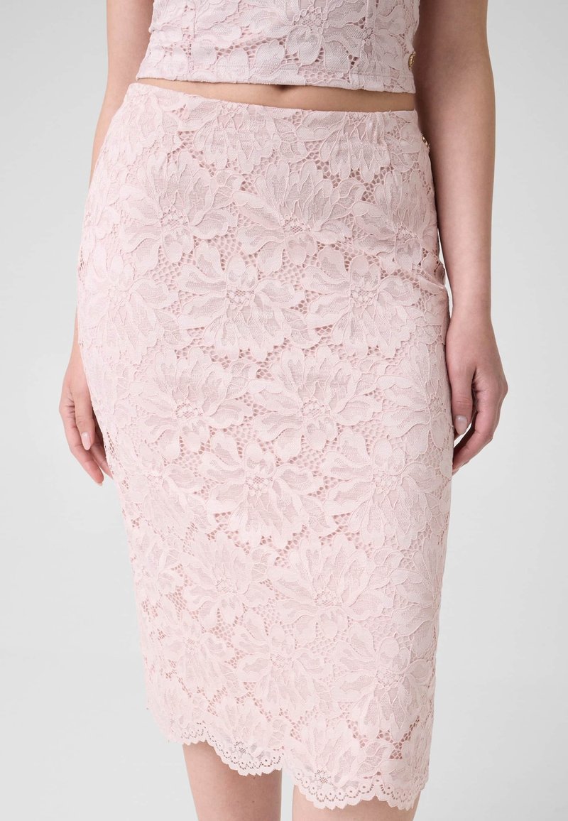 Femme portant une jupe crayon en dentelle florale rose clair et un crop top assorti, debout, bras détendus le long du corps.
