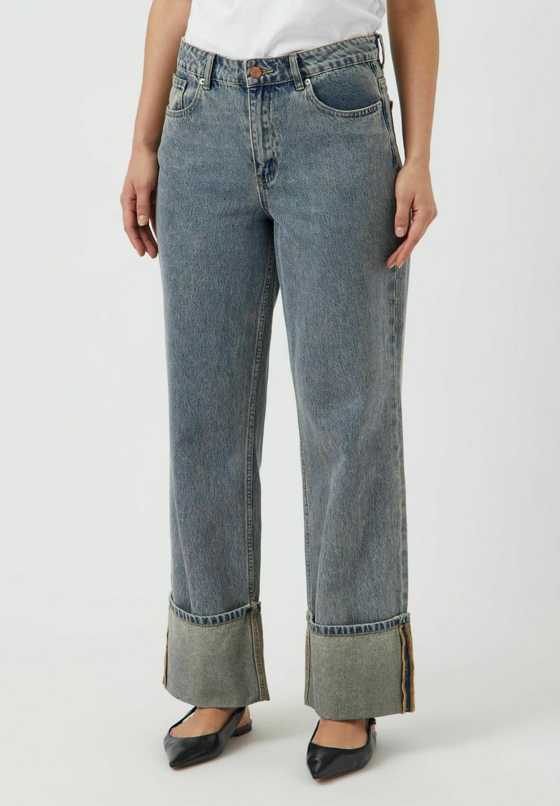 Mujer con jeans de cintura alta en azul con dobladillos anchos y zapatos planos negros de punta, de pie contra un fondo blanco liso.