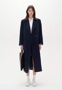 Lungo cappotto blu navy con colletto a punta e un bottone, abbinato a una camicia bianca. Il modello tiene una borsa a tracolla marrone, indossando calzini bianchi e sneakers.