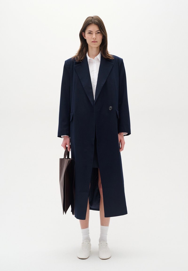 Lungo cappotto blu navy con colletto a punta e un bottone, abbinato a una camicia bianca. Il modello tiene una borsa a tracolla marrone, indossando calzini bianchi e sneakers.
