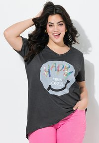 Zwarte katoenen t-shirt met een gebogen zoom, voorzien van een meerkleurige tekstgrafiek die "HAPPY VIBES ONLY" leest op een lichtgrijze smiley. 
