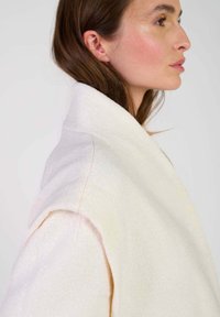 Crème wollen jas met een brede kraag en open voorkant, met een zachte textuur en minimale stiknaad details langs de randen.