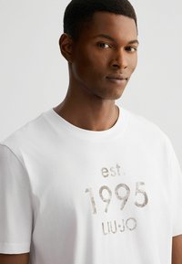 Witte katoenen T-shirt met een gestructureerde print met "est. 1995 LIU JO" in grijs. Klassieke ronde hals en korte mouwen.