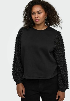 Femme portant un haut noir à manches longues avec des manches texturées à motif floral et un pantalon noir, debout devant un fond clair uni.