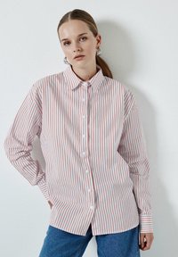 Camicia bianca con sottili righe rosse verticali, maniche lunghe, colletto classico e vestibilità rilassata, abbinata a jeans di denim blu.