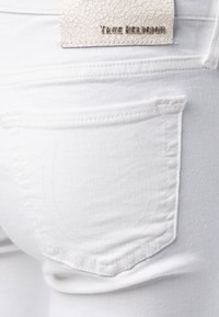 Close-up van een wit denim jeans achterzak met een gestikt hoefijzerontwerp en een gebroken leren label met de tekst True Religion.