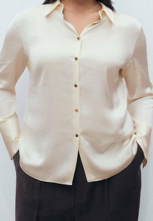 Kvinde iført en cremet satinbluse med guldknapper, stoppet ned i højtaljede mørke bukser, hænderne i lommerne.