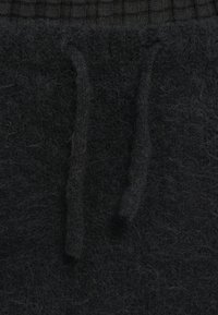 Calças pretas com cordão ajustável, feitas de material felpudo, com uma cinta texturizada que apresenta um padrão de grelha. Inclui cordões visíveis na parte da frente.