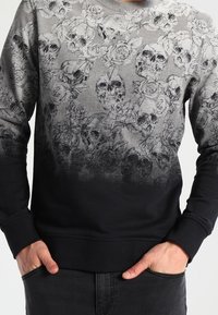 Felpa a gradiente grigio e nero con motivo dettagliato di teschio nero e rose, indossata con le mani nelle tasche anteriori.