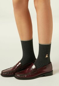 Mørkegrå ribbede sokker med guldtrim og en lille broderet bamse, båret med maroone læderloafers. Skoene har et syet design.