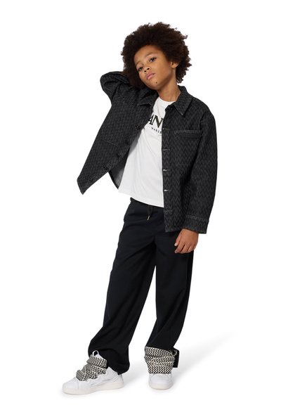 Chemise noire à motifs avec des boutons, t-shirt blanc avec logo, pantalon noir ample et baskets blanches avec des lacets à motifs noirs et blancs.