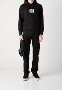 Persona vestida con una sudadera negra de Calvin Klein, pantalones negros, zapatillas negras con suelas blancas, sosteniendo un bolso cruzado negro por la correa.