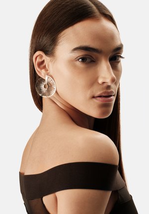 Cirkelvormige oorbellen in goudkleur met een getextureerd zonnestraalontwerp en een open midden, getoond tegen een neutrale achtergrond.