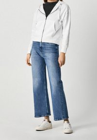 Felpa bianca con cerniera e tasche frontali, abbinata a jeans larghi blu e sneakers bianche. L'outfit presenta una vestibilità casual e rilassata.