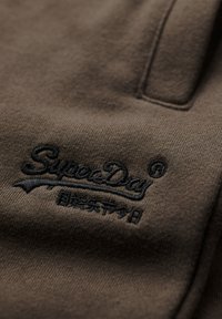 Tejido marrón con una textura suave que presenta un logo bordado en negro "SuperDay" y detalles de acento. La vista en primer plano resalta la costura y el diseño.