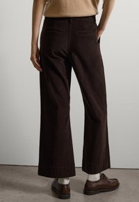 Braune Cordhosen mit weitem Bein und Gesäßtaschen, die über eine glatte Textur und einen geraden Saum verfügen, kombiniert mit dunklen Schuhen und weißen Socken.