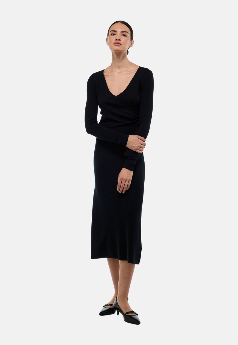 Robe tricotée noire à manches longues, décolleté en V, silhouette ajustée et longueur genou. Texture lisse, couleur unie, accents minimaux.