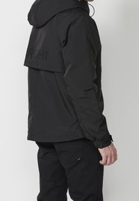 Svart belagd jacka med huva, med en kort bakdesign, justerbara ärmar och subtilt tryckt varumärke på baksidan.