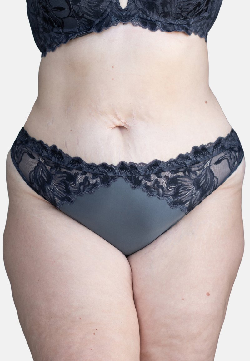 Schwarze Spitze und Netz-Bikiniunterhosen mit einem Blumenmuster entlang des Taillenbundes, einem glatten, undurchsichtigen grauen Körper und gewellten Kanten.