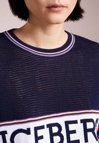 Granatowy sweter z dzianiny ma wielobarwny prążkowany kołnierz oraz wyrazisty wzór z napisem na piersi. Faktura wydaje się lekko prześwitująca.